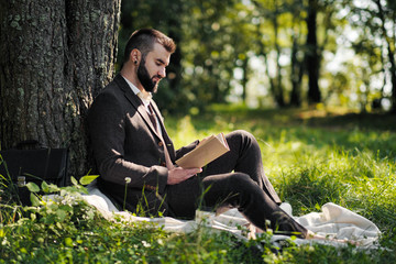 Fototapeta premium Young attractive bearded business man sitting on green grass under tree and resting in park. Read book, drink coffee. Relaxed, tired of work, lunch break.