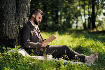 Young attractive bearded business man sitting on green grass under tree and resting in park. Read book, drink coffee. Relaxed, tired of work, lunch break.
