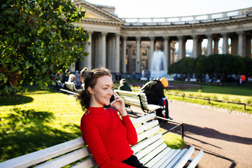 Happy smiling casual dressed female talking with friends via mobile phone while sitting outdoors near history architectural building during free time in weekend abroad. Woman having cellphone call