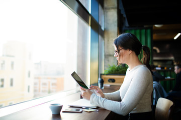 Hipster girl skilled E-book writer reading information on website via portable touch pad while sitting at workstation in coworking space. Female college student using digital gadget for education