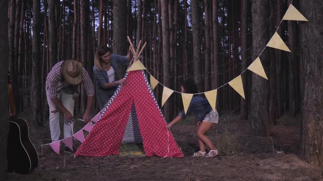 mom with dad and daughter decorate wigwam with flags and garlands between rows of trees during a forest camping trip at evening, then give each other five