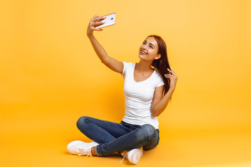 Young girl in white t-shirt sitting cross-legged and taking selfie on mobile phone, on yellow background