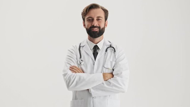 Attractive young bearded man doctor in white professional coat with stethoscope listening someone attentively and shaking his head positively while looking at the camera over gray background isolated