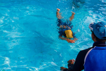 Asian little boy age 5 years old learning basic to swim in the lesson of lying and floating technique on surface water with adult male swimmer and trainer in blue swimming pool background.