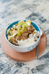 Greek yogurt, granola, kiwi and banana in bowl with a spoon on a gray concrete background, wooden tray, vertical format. Fitness diet for weight loss, proper and tasty food