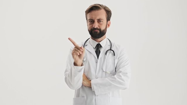 Angry Young Bearded Man Doctor In White Professional Coat With Stethoscope Saying No With Waving Finger And Shaking His Head Negatively While Looking At The Camera Over Gray Background Isolated