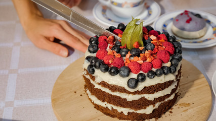 Woman cuts a piece of birthday homemade chocolate cake with cream and sponge cakes layers decorated fresh berries, closeup.