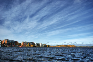 Fototapeta na wymiar View from the pier for boats on the city and lake. Outside is a cold sunny day. In the background is an island with a forest. There are residential buildings on the shore. Scandinavian lifestyle.