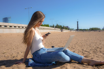Woman university student using messenger on mobile phone during online learning on laptop computer relaxing on the coastline in sunny day. Blogger chatting on cellphone while resting outdoors
