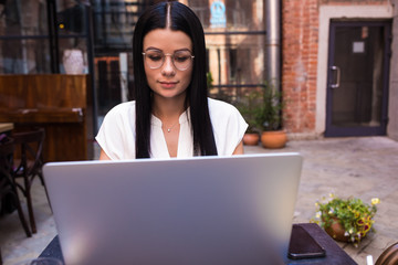 Woman skilled copywriter using applications on laptop computer, sitting in coffee shop outdoors. Female in fashionable glasses checking e-mail on notebook gadget,resting in cafe outside