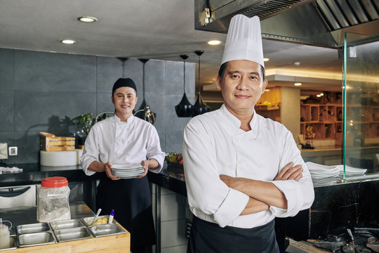 Portrait of confident Asian chef standing with arms crossed and looking at camera with young cook preparing food in the background in the kitchen - Powered by Adobe