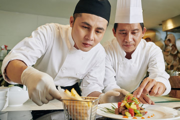 Young Asian cook decorating prepared salad together with chef in the kitchen of the restaurant