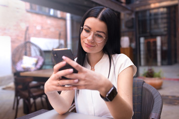Charming female with good mood received notifications on mobile phone while sitting in coffee shop. Pretty woman reading pleasant text message on cellphone while relaxing in restaurant