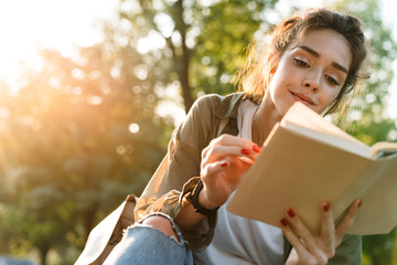 Image of young woman smiling and reading book in green park - obrazy, fototapety, plakaty