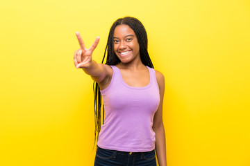 African American teenager girl with long braided hair over isolated yellow wall smiling and showing victory sign