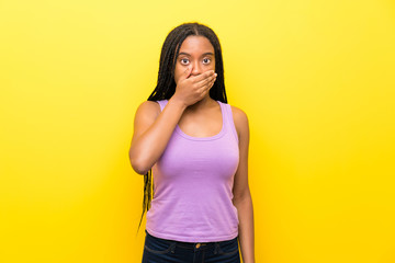 African American teenager girl with long braided hair over isolated yellow wall covering mouth with...
