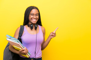 African American teenager student girl with long braided hair over isolated yellow wall pointing finger to the side - obrazy, fototapety, plakaty