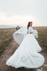 Fototapeta na wymiar Woman in fashionable dress waving veil wind. Stylish and beautiful. Bridal bouquet of orchids. Loving wedding couple outdoor. Beautiful woman with professional make up and hairstyle.