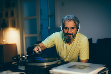 middle aged man sitting sofa and playing music on record player in his home. night scene