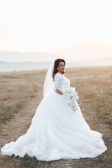 Fototapeta na wymiar Happy bride in wedding dress posing on the road in a field at sunset. Beautiful young bride is holding a wonderful bouquet of orchid. Beautiful woman with professional make up and hairstyle.