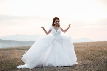 Fototapeta na wymiar Happy bride in wedding dress posing on the road in a field at sunset. Beautiful woman. Bride at wedding day walking Outdoors on nature. Beautiful and stylish bride in wedding day