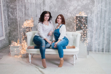 Obraz premium Two beautiful girls celebrates Christmas, New Year. Girls in the New Year's interior of the room are sitting on a white sofa against the background of garlands