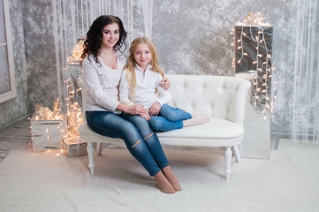 Two very pretty girls, sisters celebrates Christmas, new year. Girls in the New Year's interior of the room are sitting on a white sofa against the background of garlands