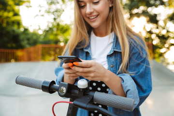 Smiling cheerful teenage girl in park walking on scooter using mobile phone.