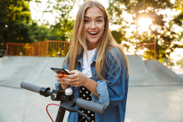 Smiling cheerful teenage girl in park walking on scooter using mobile phone.