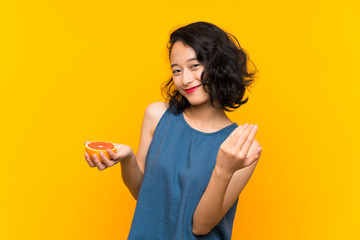 Young asian girl holding a grapefruit over isolated orange background inviting to come with hand. Happy that you came