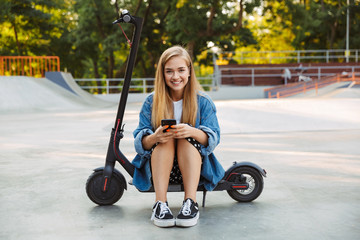 Happy teenage girl in park sit on scooter using mobile phone.
