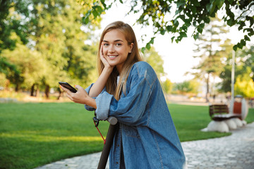 Cute young teenage girl outside in nature green park with scooter using mobile phone.