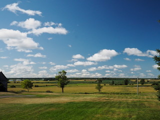 Blue sky and beautiful clouds above multi-colored farm fields of colorful agricultural crops, trees and barn. Pastoral and beautiful landscape background. Copy space.