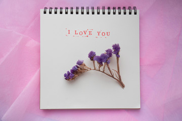 Text I love you in English, typed in stamps, on a white background, decorated with dried purple flowers. Shooting from above, flat lay, copy space. Notepad mockup with place for text.