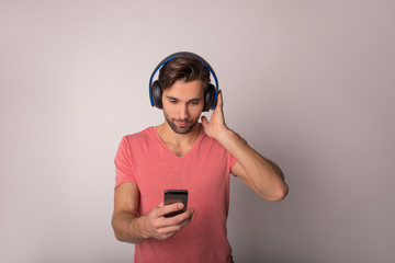 Bearded beautiful man in pink casual shirt watching video on mobile phone and using wireless headphones while standing isolated in studio against wall background with copy space