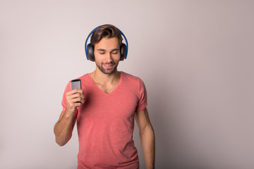 Handsome casual dressed hipster guy enjoying while listening cool music via  via wireless headphones and mobile phone while standing isolated in studio.