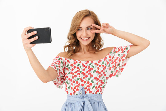 Portrait of positive young woman smiling and showing peace fingers while taking selfie photo on cellphone