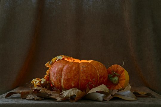 A Ripe Orange Pumpkin, With Light From A Candle Inside, With Seeds Hanging From The Inside Out And A Nearby Cut Out Hat Cap With A Green Handle. On A Velvet Brown Background Among Dry Autumn Leaves.