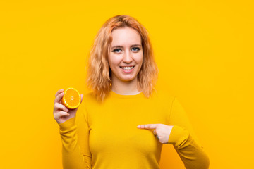 Young woman holding an orange over isolated yellow background with surprise facial expression