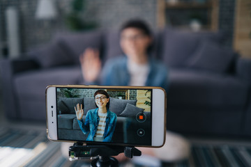 Joyful girl recording video with smartphone camera talking waving hand at home, focus on mobile...