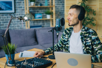 Joyful guy is talking in microphone wearing headphones using laptop in studio sitting at desk alone...