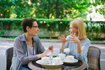 Beautiful mature ladies friends are talking sitting in street cafe together enjoying conversation. People, modern lifestyle and female friendship concept.