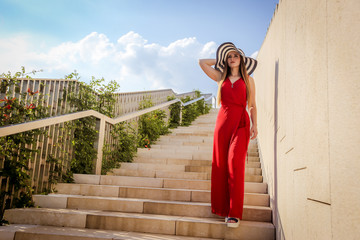 A girl in a summer hat in sunglasses in a red overalls stands on the stairs in the park. City fashion.