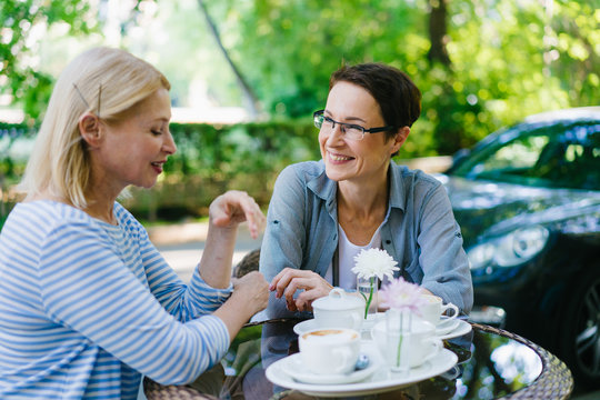 Beautiful mature woman is talking to female friend in street cafe smiling having fun enjoying warm summer day. Modern lifestyle, friendship and conversation concept.