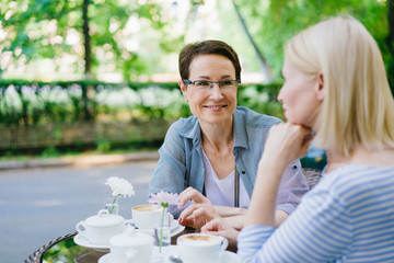 Attractive adult women friends blonde and brunette are speaking in outdoor cafe sitting at table with drinks. Conversation, friendship and people concept.