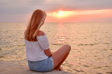 Fototapeta na wymiar A girl with blond hair is sitting on the pier watching the sunset