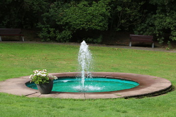 Kleiner Teich mit Springbrunnen im Park auf einer Wiese und mit Sitzbank