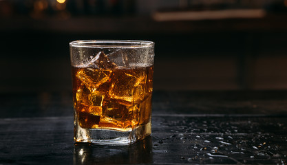 Glass of Whisky (whiskey), bourbon, ice tea or coke (cola) soda with ice and water drops on wood table at dark background with copy space