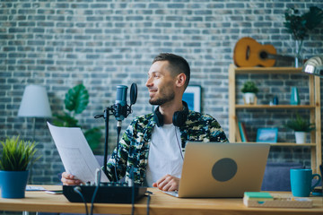Attractive guy is recording podcast using microphone holding piece of paper indoors creating...