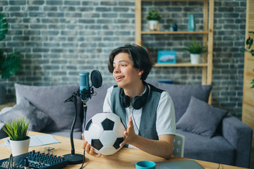 Happy teenager sports fan is talking in microphone in recording studio holding football creating...